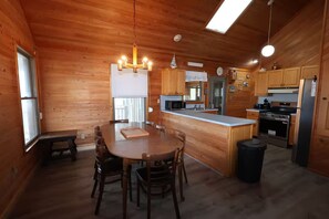 A wooden cabin interior with a dining table, chairs, a kitchen with appliances, and a large window. - 4 Bedroom Oceanfront Home in Kitty Hawk, NC. JAX’S PLACE (Kitty Hawk)