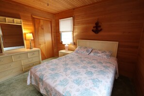 A wooden-paneled room with a bed, a dresser, a nightstand, a lamp, and a window with blinds. - 4 Bedroom Oceanfront Home in Kitty Hawk, NC. JAX’S PLACE (Kitty Hawk)