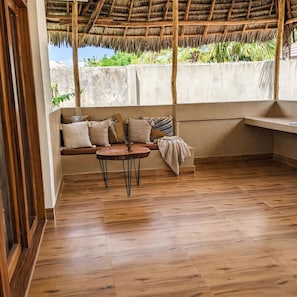 Quarto com piso de madeira, telhado de palha, um banco com almofadas e uma pequena mesa.