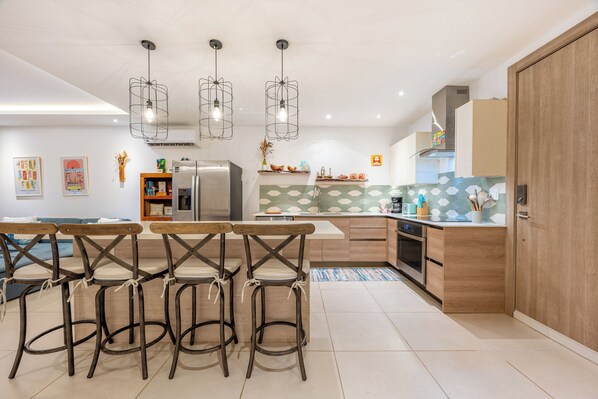 A modern kitchen with a central island, bar stools, a refrigerator, and pendant lighting. - Tamarindo Ocean-View Condo, Shared Infinity Pool (Tamarindo)