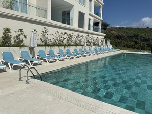 A poolside area with lounge chairs and a clear pool, surrounded by a building with balconies. - Tamarindo Ocean-View Condo, Shared Infinity Pool (Tamarindo)