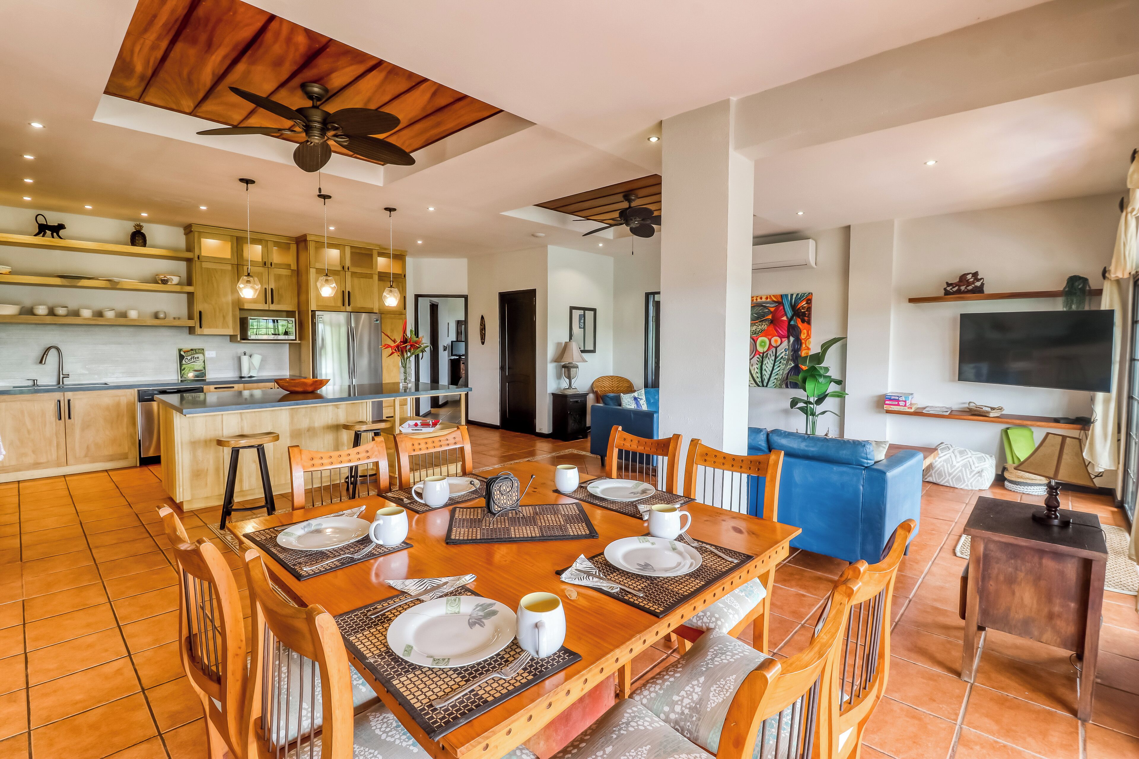 Cozinha moderna com armários de madeira, área de jantar com mesa de madeira posta para quatro pessoas e sala de estar com sofá azul e televisão de tela plana.
