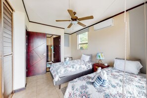 Ein Hotelzimmer mit zwei Betten, einem Deckenventilator, einer Klimaanlage, einem Fenster mit Blick und einer Tür zu einem anderen Raum.