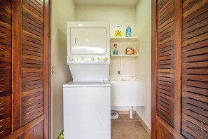 A compact laundry room with a washing machine and dryer, wooden shutters, and a shelf with cleaning supplies. - Stunning Lake Arenal Views: Duplex Unit w/ Patio! (La Fortuna)
