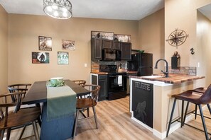 A modern kitchen with dark cabinets, a central island, and a dining area with wooden chairs and a table. - Bigfoot’s Bunk: Cozy Cabin on Table Rock Lake (Kimberling City)