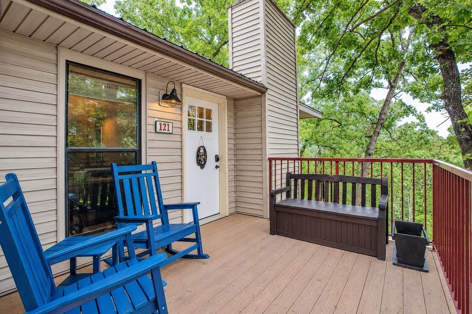 A wooden deck with a bench and a table, a white door with a number sign, and a red railing.