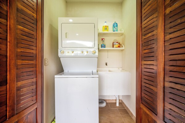 A compact laundry room with a washer and dryer, a small shelf with cleaning supplies, and wooden slatted doors. - Near Lake Arenal & Volcano: Duplex Unit w/ Views! (La Fortuna)