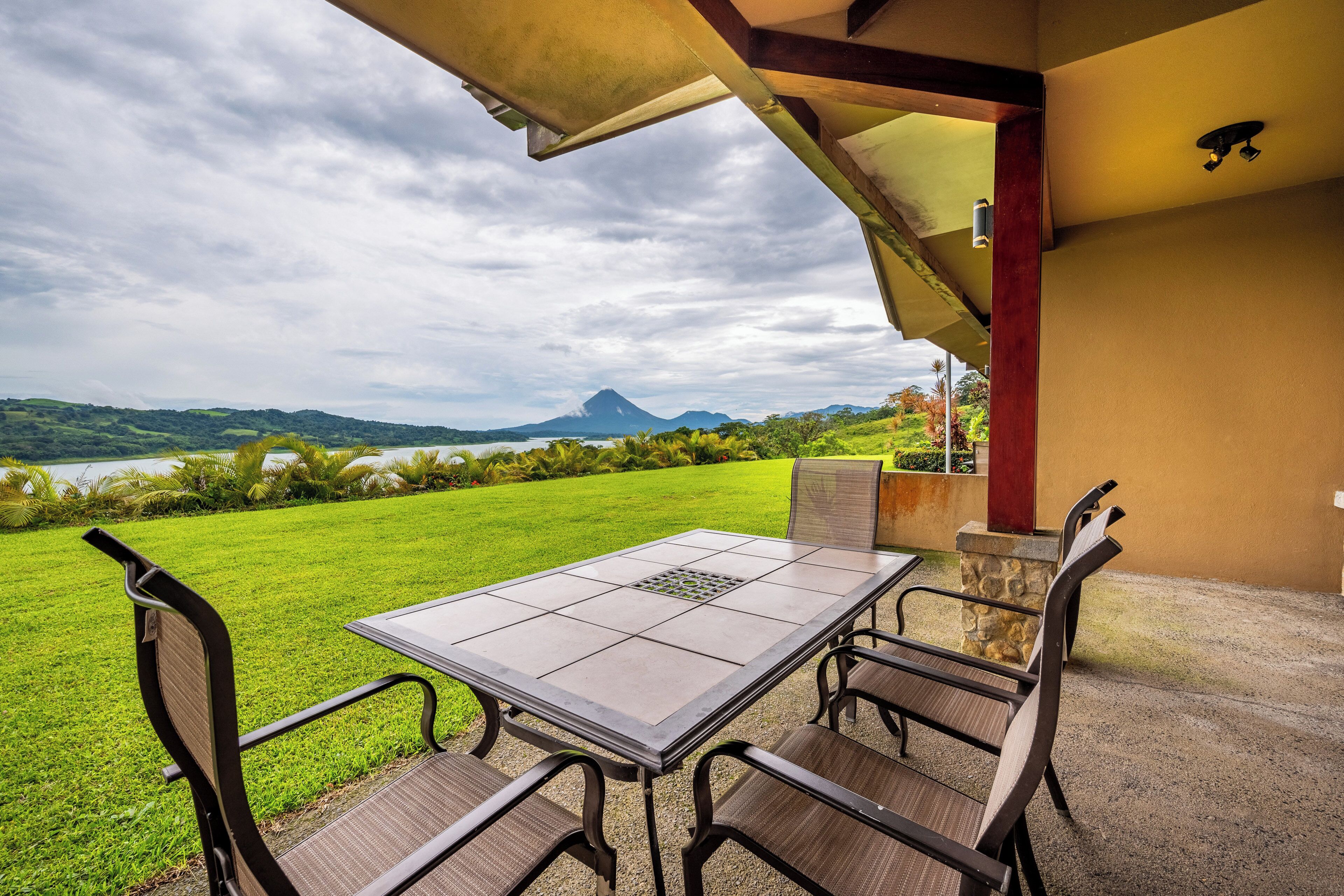 Un patio exterior con una mesa y sillas, con vista a un área verde con una montaña al fondo.