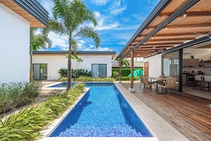 A modern outdoor pool area with a wooden deck, a dining area with chairs, and a kitchen visible through a glass door. - Walk to Beach! Tamarindo Home w/ Private Pool (Tamarindo)