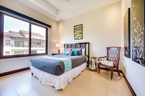 A bedroom with a large bed, a chair, a nightstand, and a window with a view of buildings. - Playa Hermosa Penthouse: Ocean Views, 2 Pools (Playa Hermosa)