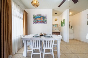 A dining area with a white table set for four, white chairs, a painting of a leopard on a sofa, and a kitchen in the background. - Playas del Coco Condo w/ Pool: Walk to Beach! (Playas del Coco)