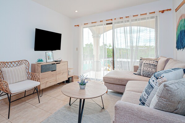 A modern living room with a sofa, chairs, a coffee table, a TV, and a view through a glass door. - 3 Mi to Beach: Modern Santa Cruz 2BR Condo (Tamarindo)