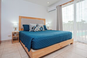 A bedroom with a large bed, two bedside tables, a nightstand, a window with curtains, and an air conditioning unit. - 3 Mi to Beach: Modern Santa Cruz 2BR Condo (Tamarindo)