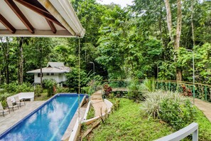 Une piscine avec une terrasse, entourée d’une végétation luxuriante et une maison en arrière-plan.