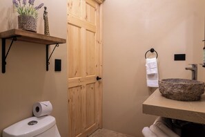 A bathroom with a wooden door, a toilet, a shelf with decorative items, a towel rack, and a stone sink. - Uvita Studio 2 Mi to Beach: Fast WiFi (Uvita)