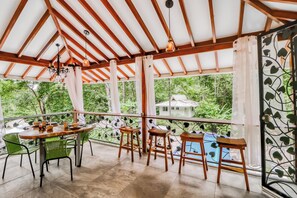 A covered outdoor area with a wooden table and chairs, a chandelier, and a view of greenery.