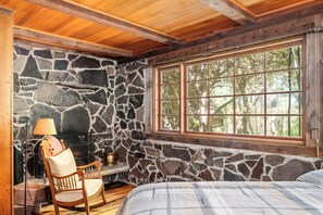 Una habitación rústica con pared de piedra, techo de madera, un ventanal grande, una cama, un sillón y chimenea.