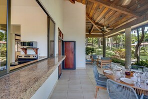 A modern kitchen with a built-in microwave and stove, a dining area with a table set for four, and a view of a lush garden. - Potrero Beachfront: Ocean-View Patio, W/D! (Playa Penca)