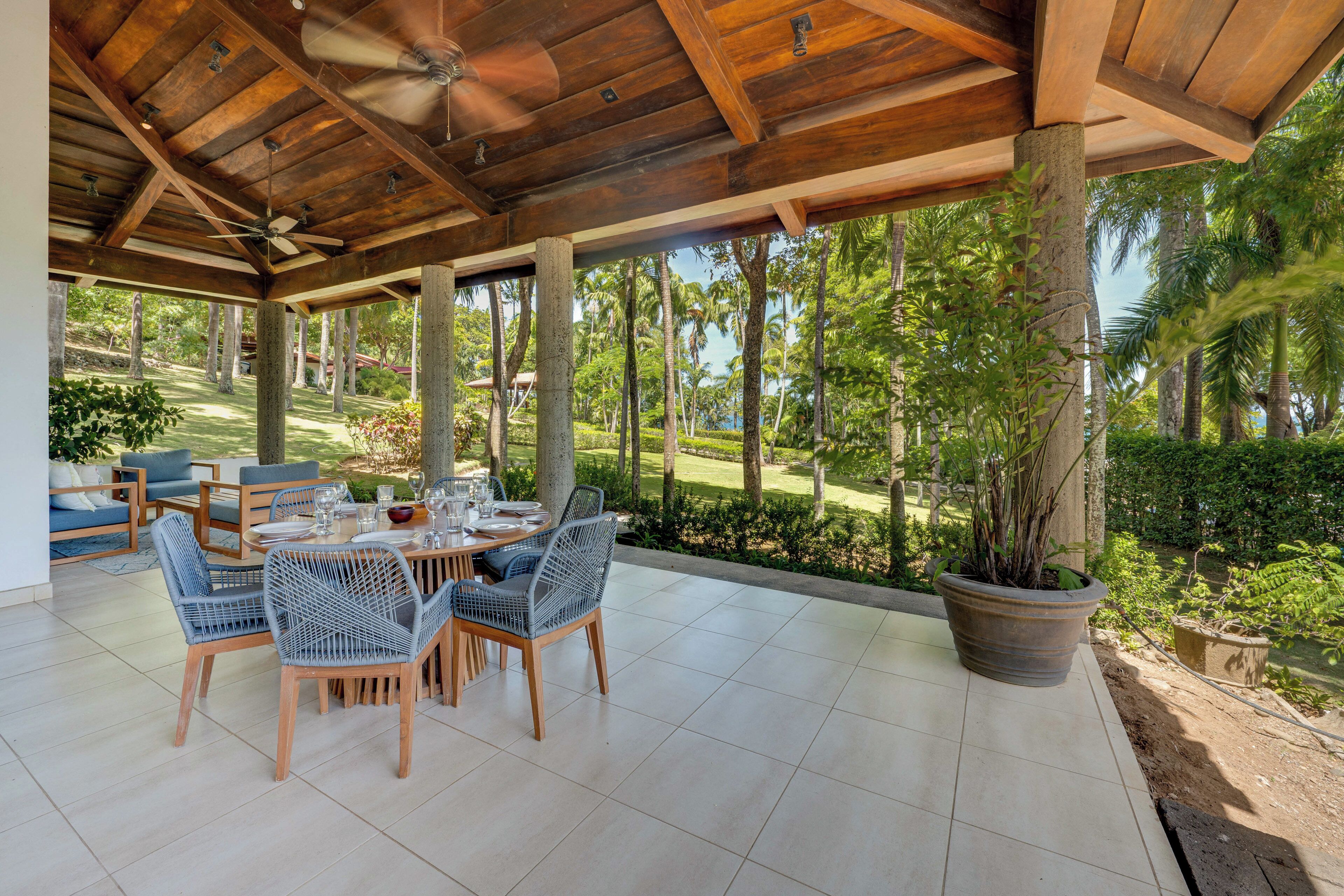 A covered outdoor dining area with a table set for four, surrounded by wicker chairs, under a wooden ceiling with a fan.