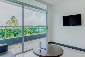 A hotel room with a large balcony, a flat-screen TV mounted on the wall, and a round wooden table with a remote control on it.
