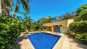 Piscina com borda azulejada, cercada por vegetação e um edifício com porta e janelas.