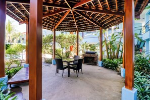 Un espace extérieur aménagé, abrité et doté de tables et de chaises, entouré de plantes et offrant une vue sur les bâtiments.