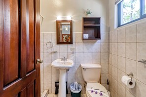 Une salle de bain avec une porte en bois, un lavabo blanc, des toilettes, une petite étagère et un miroir.