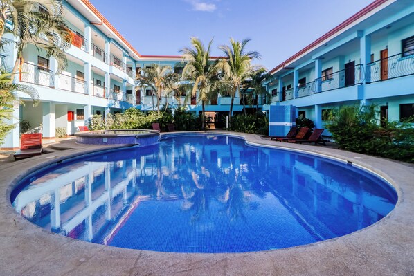 Une piscine entourée d’un immeuble à plusieurs étages, doté de balcons, de palmiers et de chaises longues.