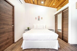 Een slaapkamer met een houten plafond, een bed met wit beddengoed, een nachtkastje met een plant en een garderobe met spiegel.