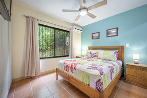 Ein Schlafzimmer mit einem Bett, einem Nachttisch, einem Deckenventilator, einer Klimaanlage, einem Fenster mit Vorhängen und Blick auf Grünfläche vor dem Fenster.