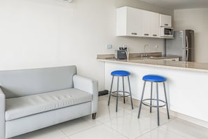 A compact kitchen with a white countertop, a built-in microwave, a stainless steel refrigerator, and a grey sofa.