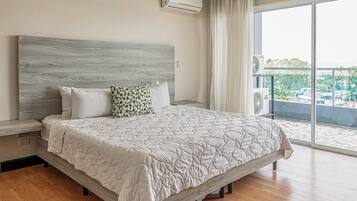 A bedroom with a large bed, a headboard, an air conditioning unit, a window with curtains, and a view of greenery outside.