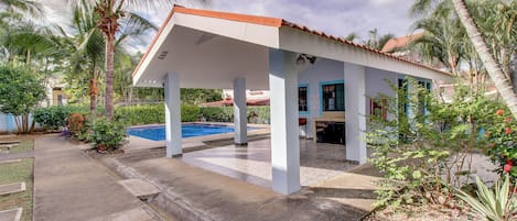 A covered outdoor area with a pool, palm trees, and a building with a tiled roof.