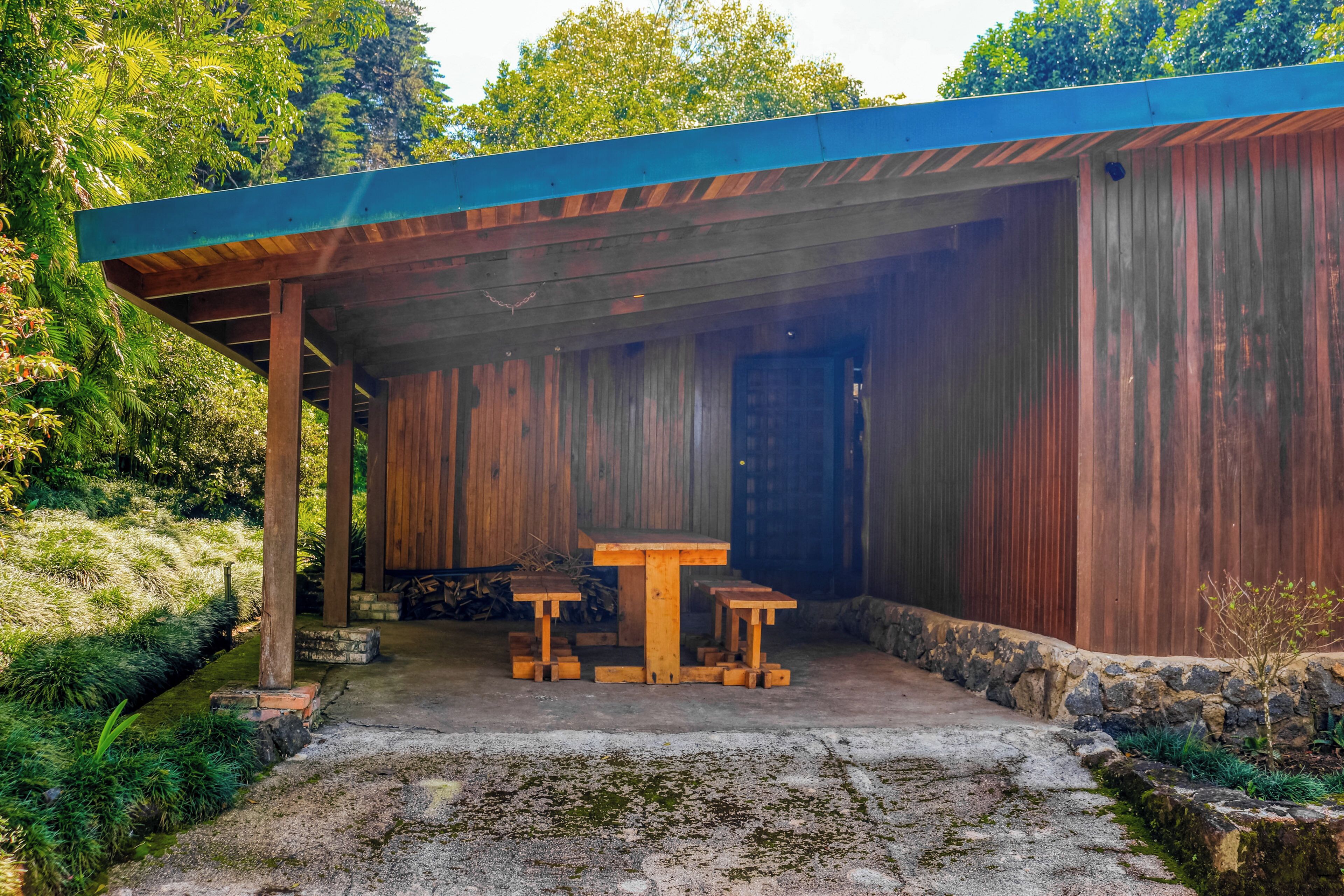 Un espacio exterior cubierto con una mesa y bancos, rodeado de follaje y un muro de piedra.