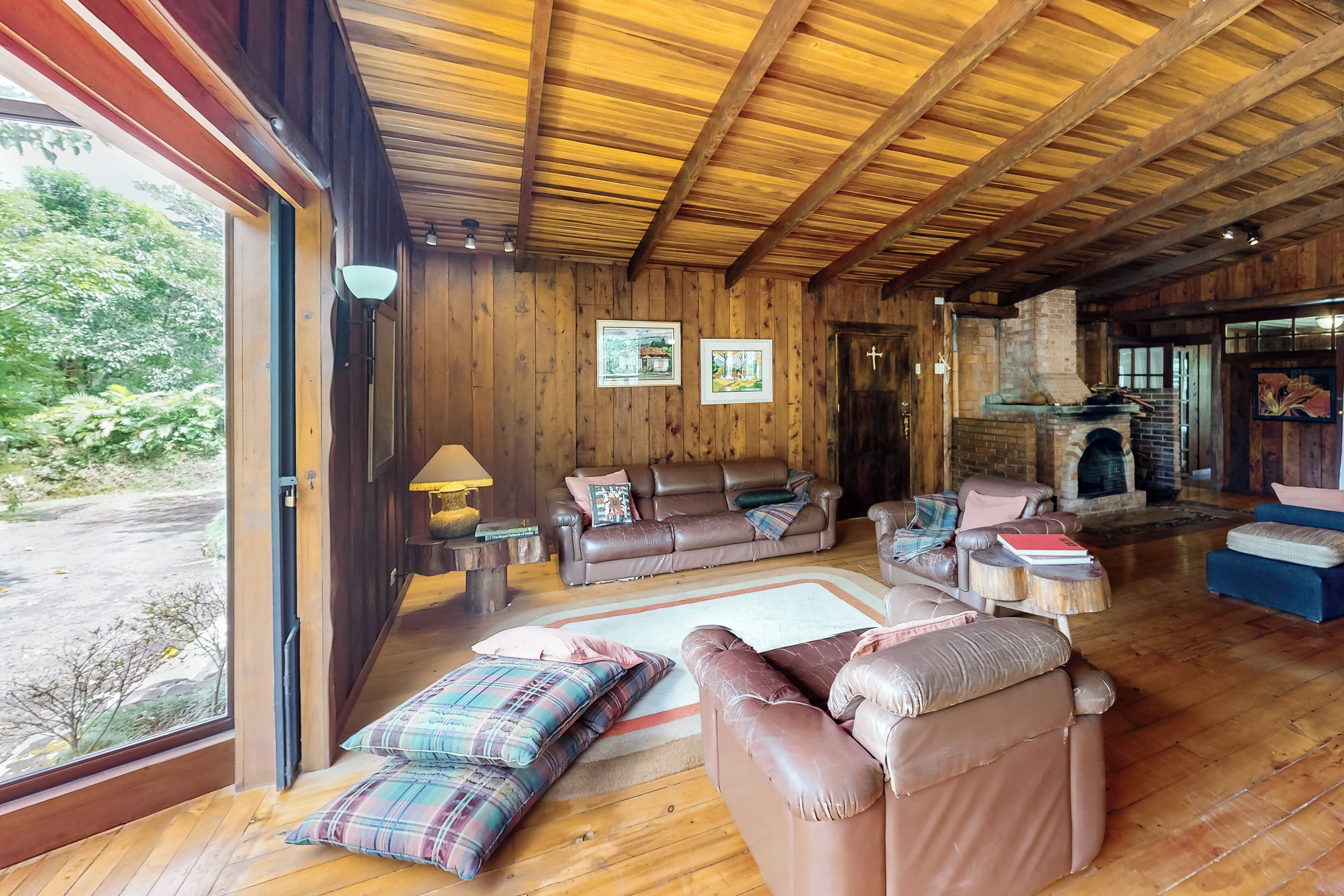 A cozy living room with wooden walls and ceiling, a fireplace, and a large window overlooking greenery.