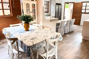 A dining area with a table set for four, white chairs, a potted plant, and a kitchen area with a refrigerator and sink in the background.