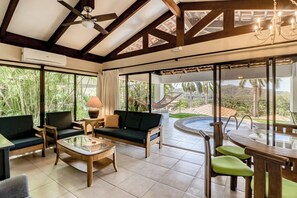 A spacious living area with a large window, a ceiling fan, a sofa, a coffee table, and a view of a swimming pool and greenery. - Beach Access & Resort Perks: Playa Hermana Villa (Playa Hermosa)
