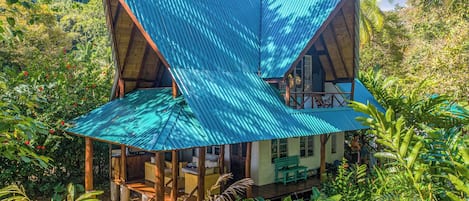 Une maison à deux étages, avec un toit bleu ondulé, entourée d’une végétation luxuriante.
