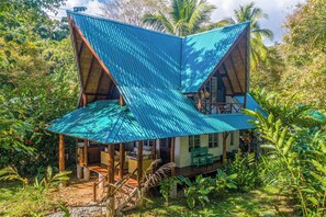 Une maison à deux étages, avec un toit bleu ondulé, entourée d’une végétation luxuriante.