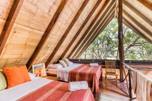 Ein Zimmer mit schrägen Wänden, Holzbalken, einem Bett mit rot-weiß gestreifter Bettdecke, einem Holzstuhl und Blick auf Bäume.