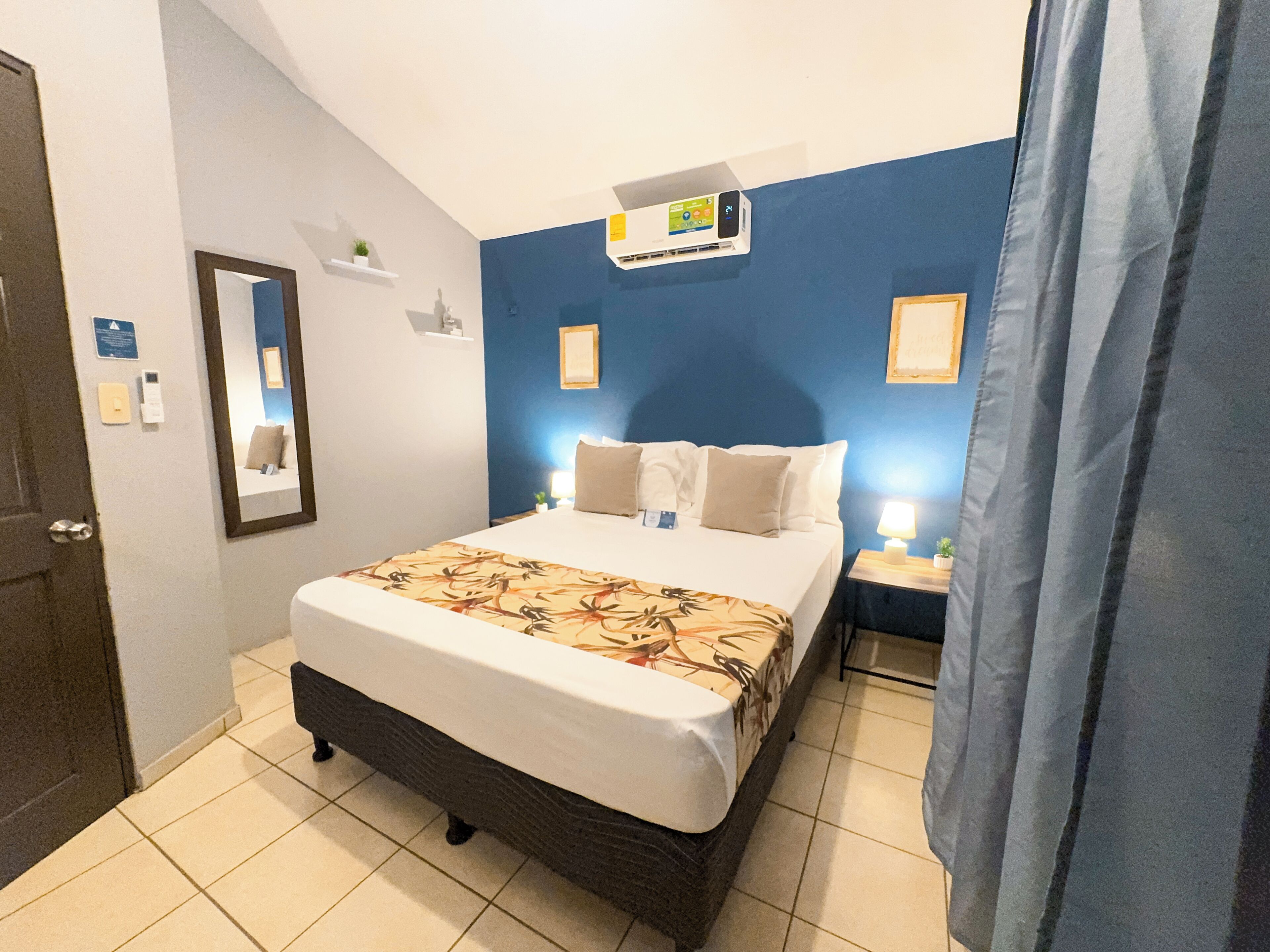 Une chambre d’hôtel comprenant un lit, des tables de chevet, un miroir, un climatiseur fixé au mur et une porte.