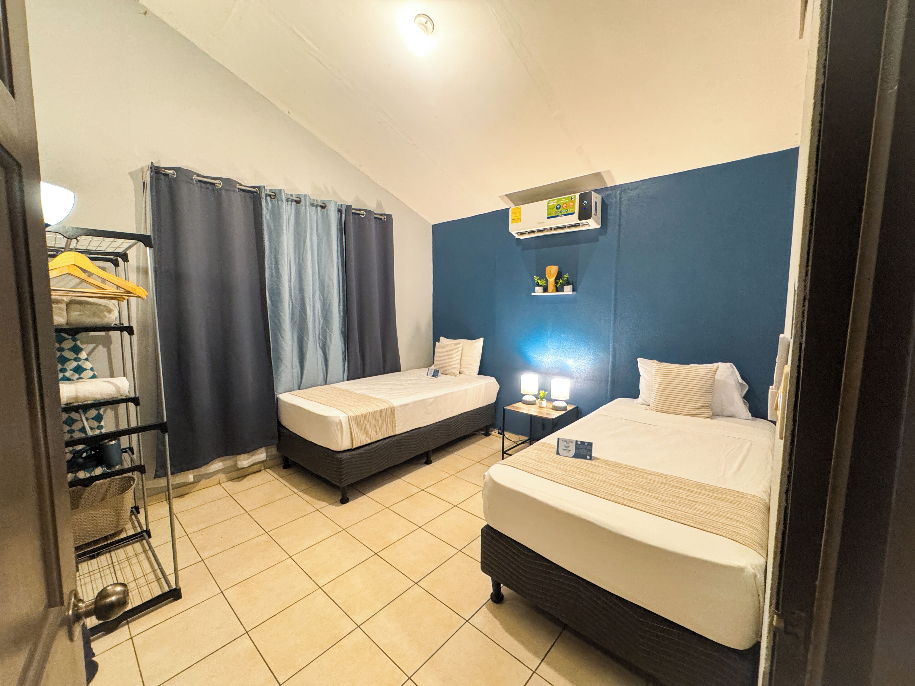 Une chambre d’hôtel avec deux lits simples, une petite table de chevet avec une lampe, un climatiseur fixé au mur et une étagère avec des articles de toilette.