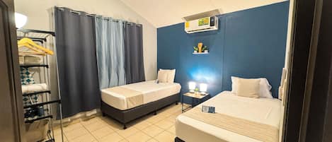 A hotel room with two single beds, a small bedside table with a lamp, a wall-mounted air conditioner, and a shelf with toiletries.
