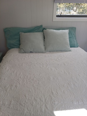 A bed with a patterned quilt and two pillows, a window with a view of trees, and a white wall.