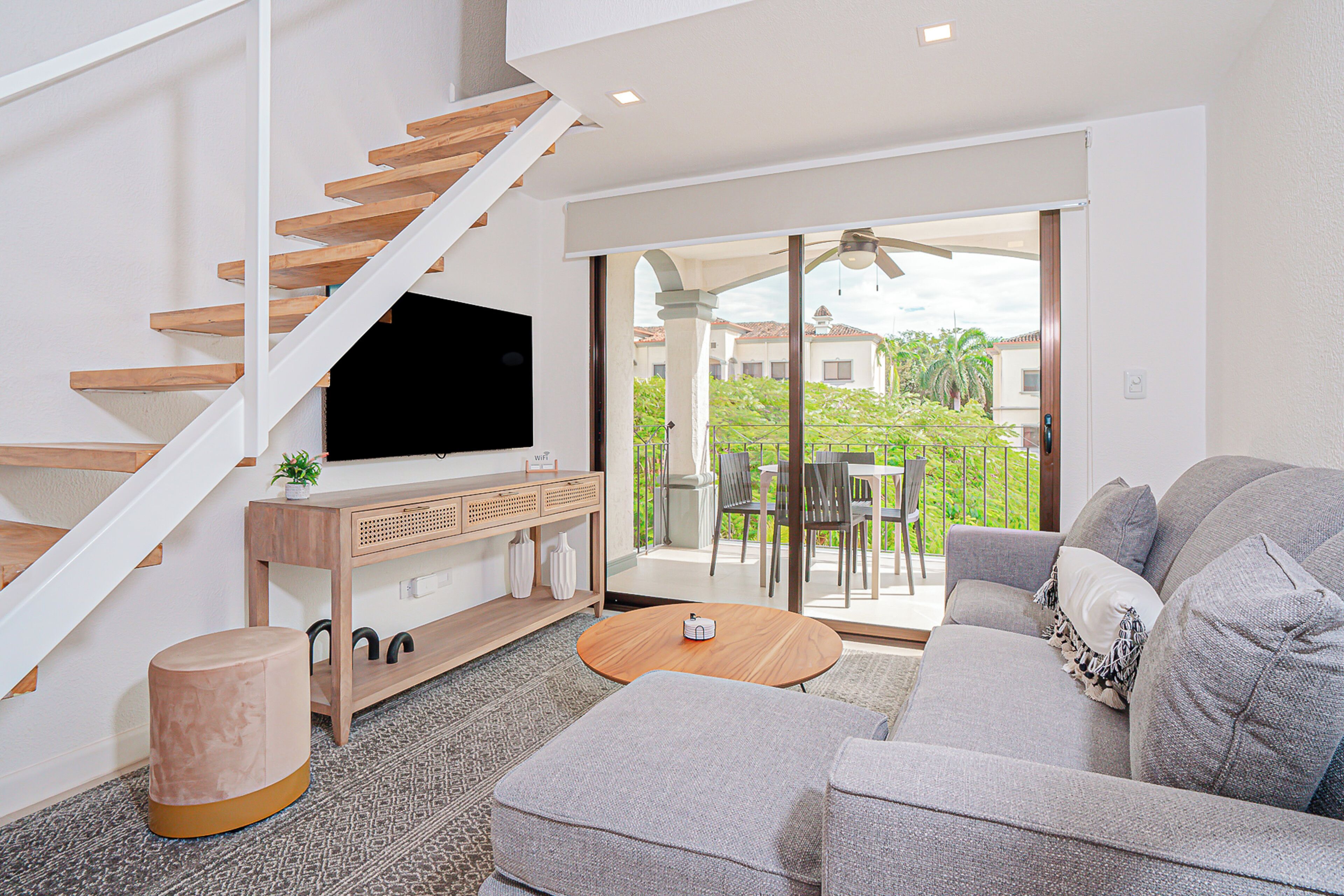 Un soggiorno moderno con una scala, una televisione a schermo piatto, un tavolino in legno, un divano grigio e un balcone con mobili da esterno.