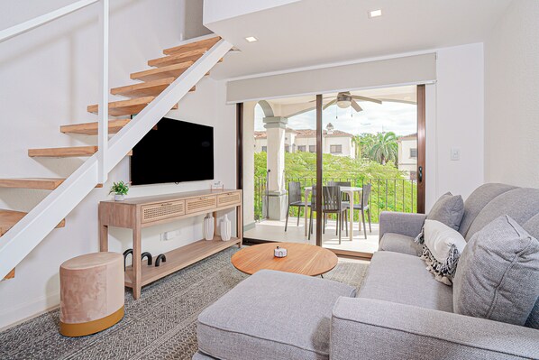 Een moderne woonkamer met een trap, een flatscreen televisie, een houten salontafel, een grijze bank en een balkon met tuinmeubilair.