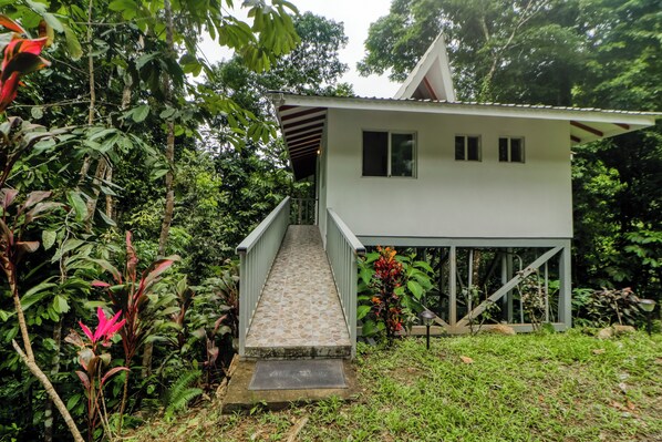 Un bâtiment à deux étages, doté d’un escalier menant à un balcon, le tout entouré d’une végétation luxuriante.