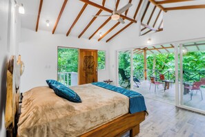 Une chambre à coucher avec un lit, une tête de lit en bois, un ventilateur de plafond et une vue sur l’extérieur grâce à de grandes fenêtres.