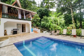 Une piscine avec un bord carrelé, un barbecue et un espace extérieur couvert offrant une vue sur la végétation environnante.