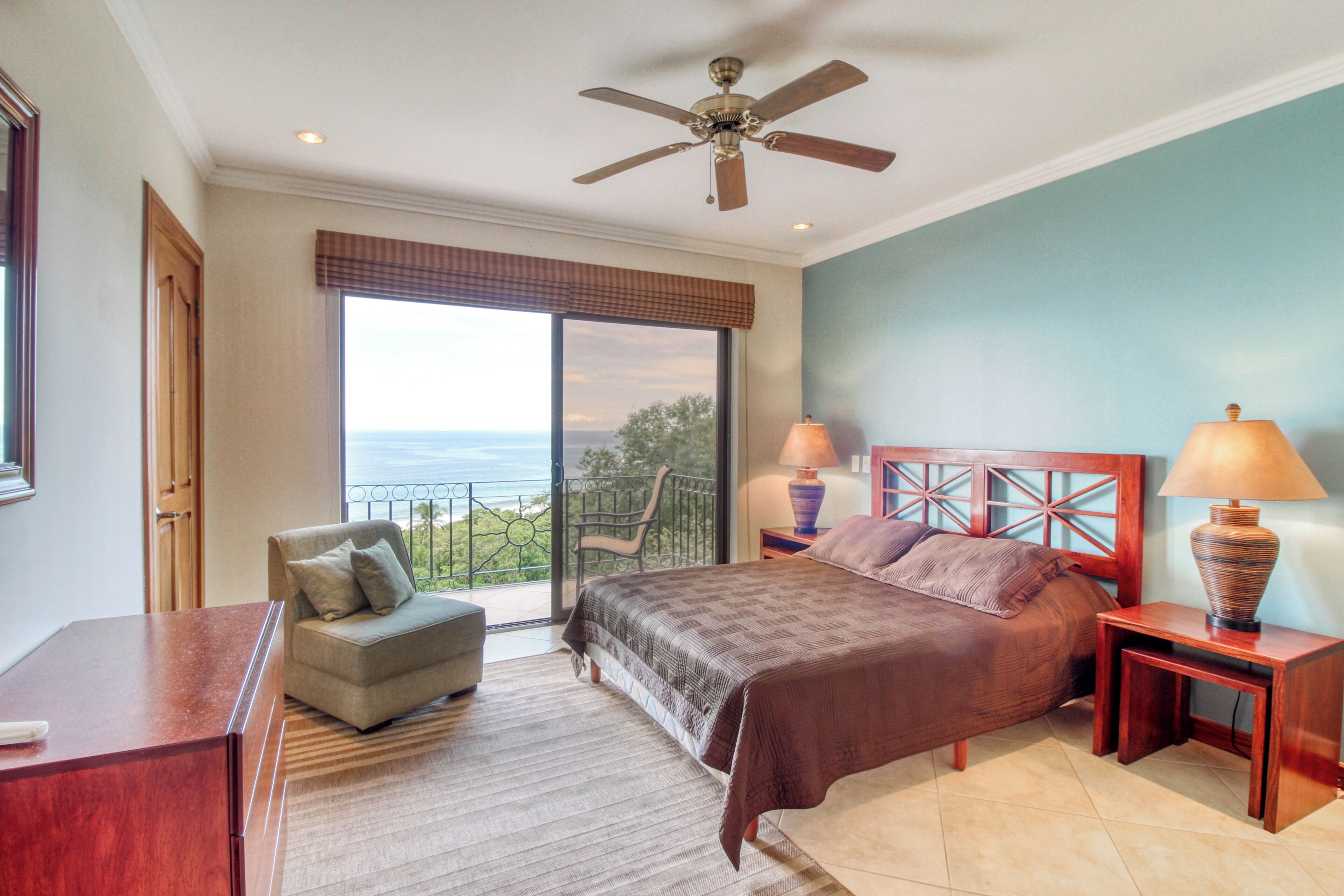 A bedroom with a bed, a chair, a desk, a ceiling fan, and a view of the ocean.